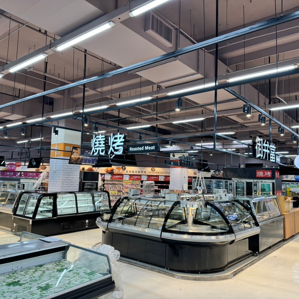 LED 連鎖通路照明|高光效線性燈與軌道燈設計 連鎖大賣場熟食與烘焙區,天花板吊掛 LED 線性燈與軌道投射燈,燒烤與烘焙招牌清楚,冷藏展示櫃整齊排列,照明明亮均勻。