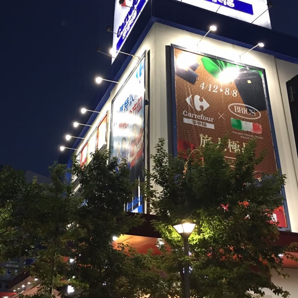 LED 外牆燈照明|建築立面與亮化工程 商場外牆的大型廣告看板,牆面排列多盞 LED 外牆投射燈向下打光,立面與行道樹清晰可見,招牌辨識度高。